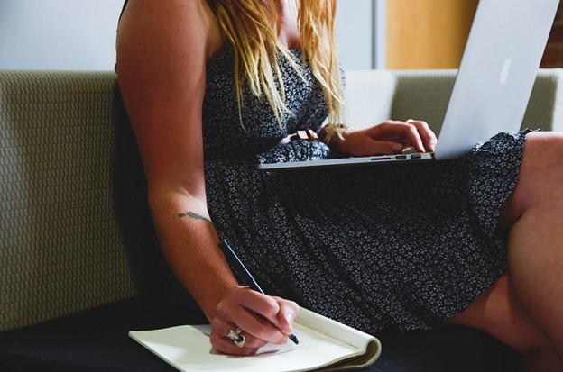 woman writing notes on a pad of paper as she is working on a laptop; writing goal to stay focused when working from home